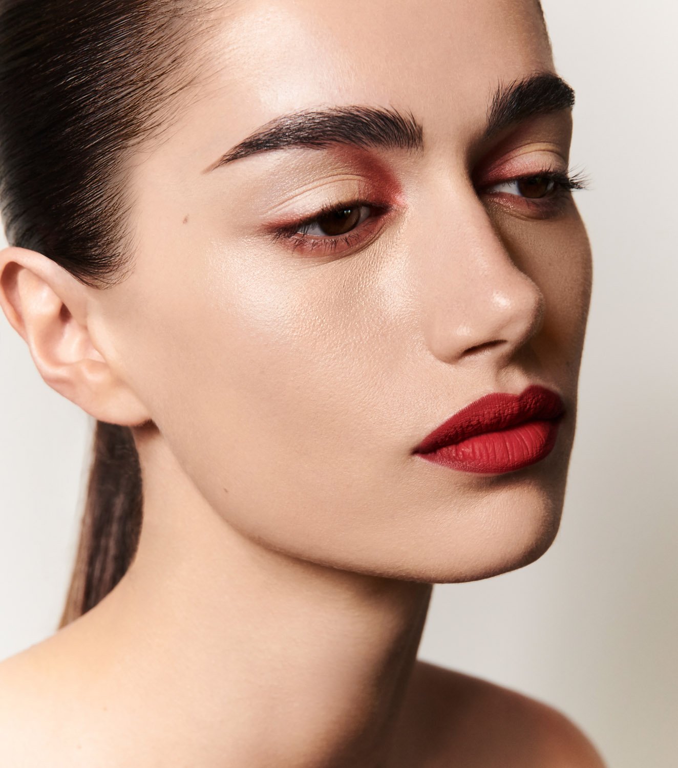 Close-up portrait of a young adult female model with red lipstick and red eyeshadow against a neutral background in a beauty setting.