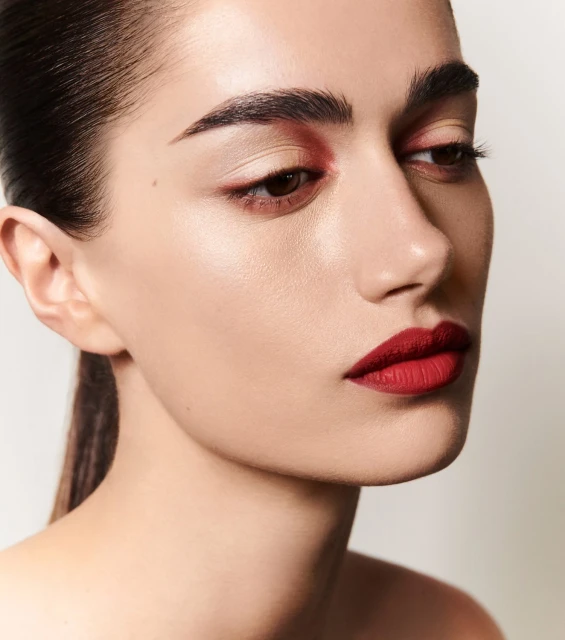 Close-up portrait of a young adult female model with red lipstick and red eyeshadow against a neutral background in a beauty setting.