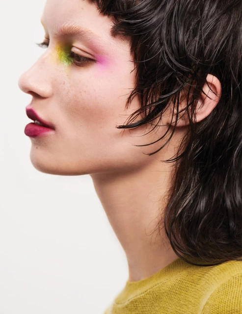 Close-up profile of a woman with vibrant makeup: yellow, green, and pink eyeshadow, dark lipstick, short dark hair, and a yellow sweater.