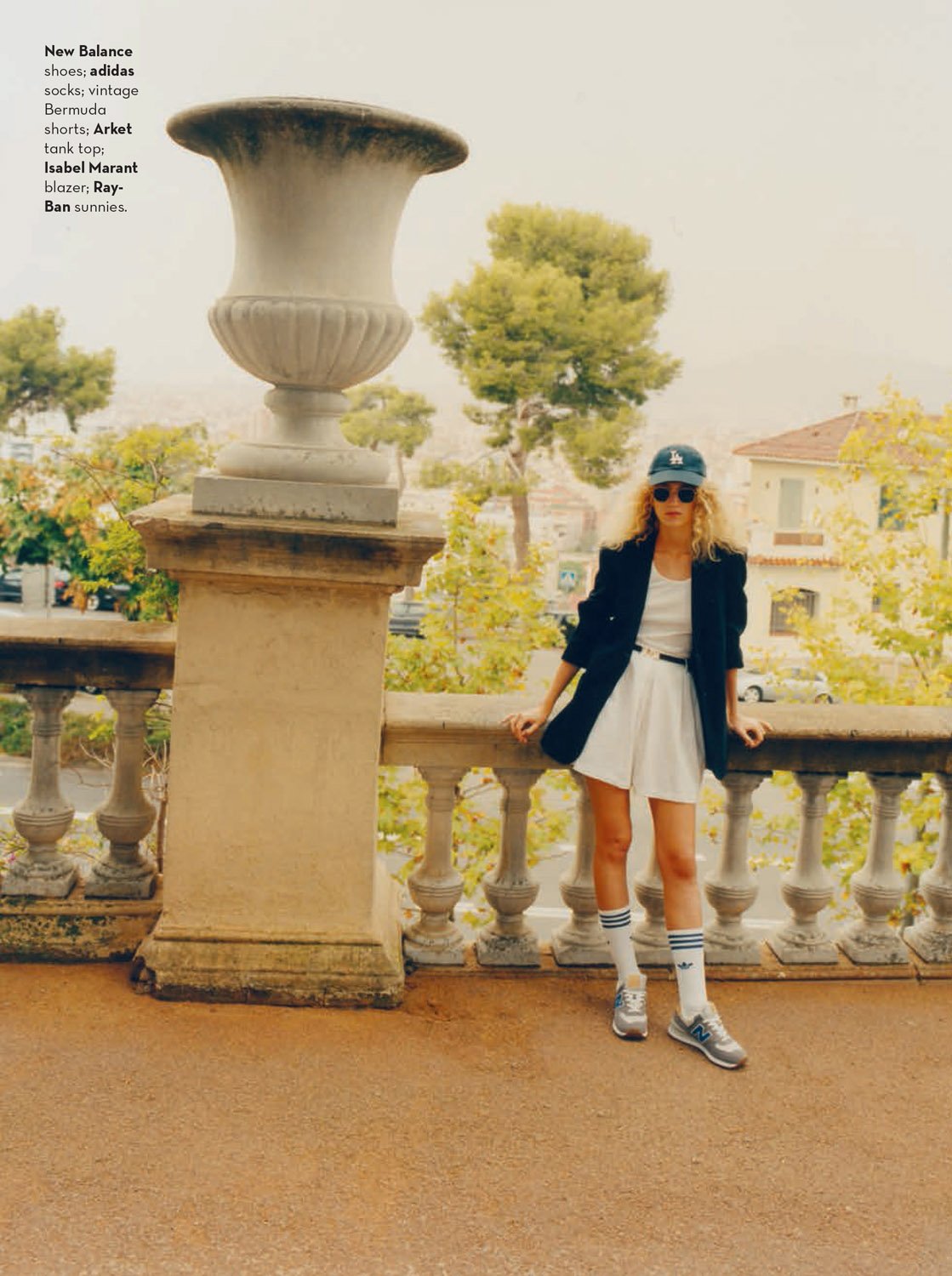 Woman in blazer, shorts, and Adidas sneakers poses on a terrace with balustrades and greenery in the background on an overcast day.