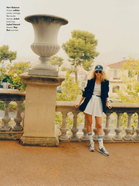 Woman in blazer, shorts, and Adidas sneakers poses on a terrace with balustrades and greenery in the background on an overcast day.