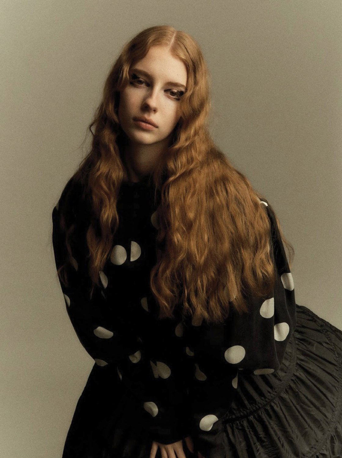 Portrait of a young woman with long red hair wearing a black, long-sleeved, polka-dot top and a black skirt against a neutral background.