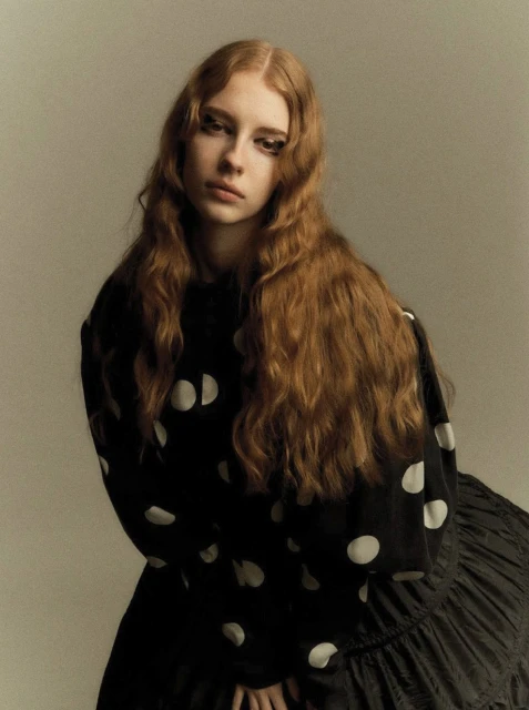 Portrait of a young woman with long red hair wearing a black, long-sleeved, polka-dot top and a black skirt against a neutral background.