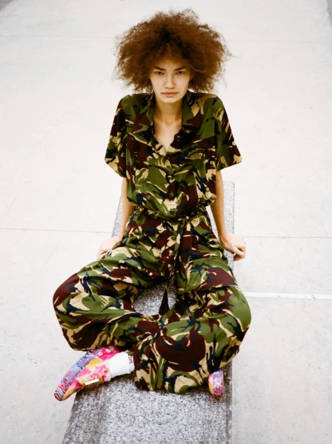 A young woman sits with legs crossed, wearing a camo jumpsuit, gazing at the camera with neutral expression. Background is a light grey.