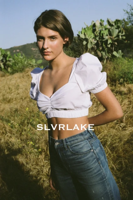 A young woman poses in jeans and a white top in a grassy outdoor setting under a sunny sky, with blurred trees in the background.