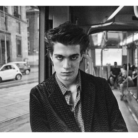 Black and white portrait of a male fashion model on a train, wearing a patterned shirt and jacket, gazing intensely at the camera.