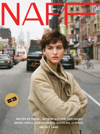 Model with short brown hair wearing a beige jacket standing on a city street with cars and buildings in the background.