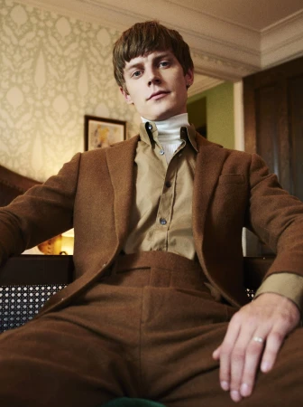 Low angle shot of a young man wearing a brown wool suit jacket, matching trousers, tan collared shirt, and a white turtleneck.