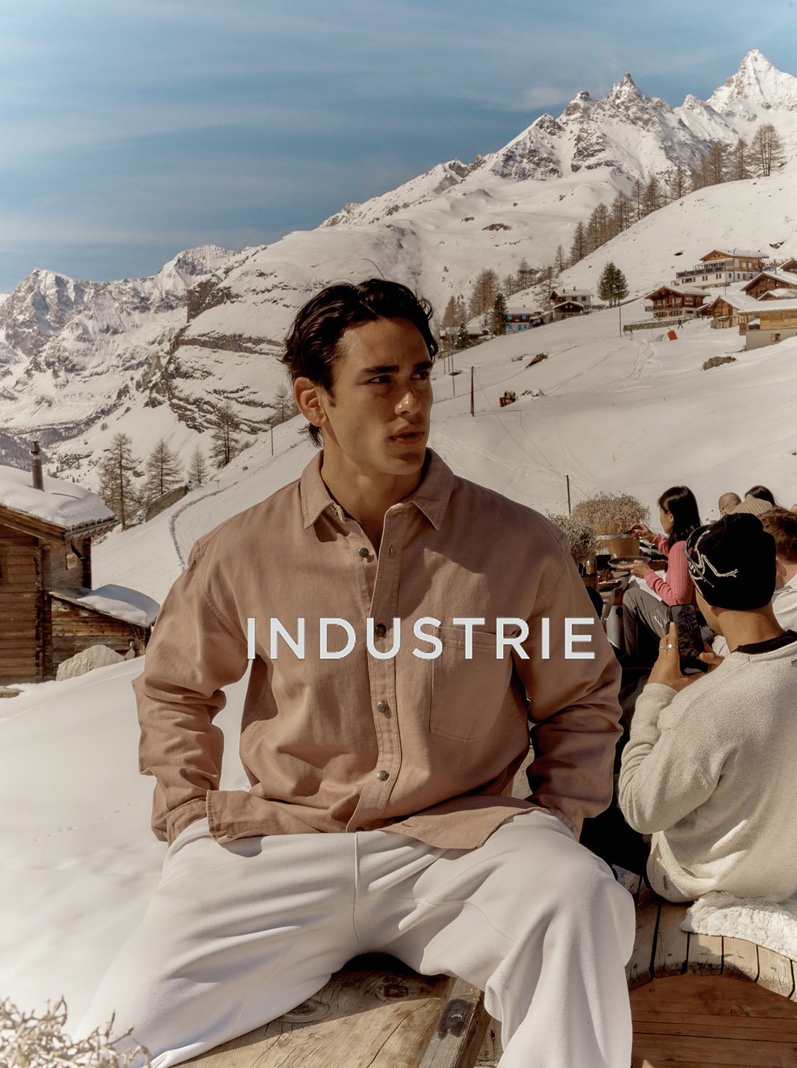 Fashion shot featuring a young adult male in an outdoor snowy mountain setting. The model wears a light brown shirt and white pants.