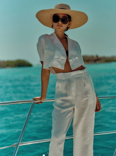 A young woman in white linen clothing and a straw hat poses on a boat with turquoise water and islands in the background, fashion shoot.