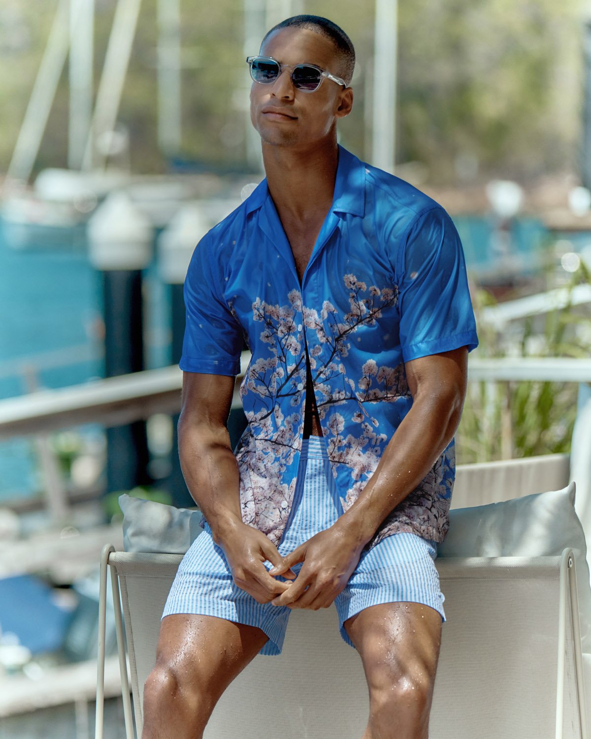 Man wearing blue floral shirt and light blue striped shorts seated outdoors near a marina by Adrian Mesko