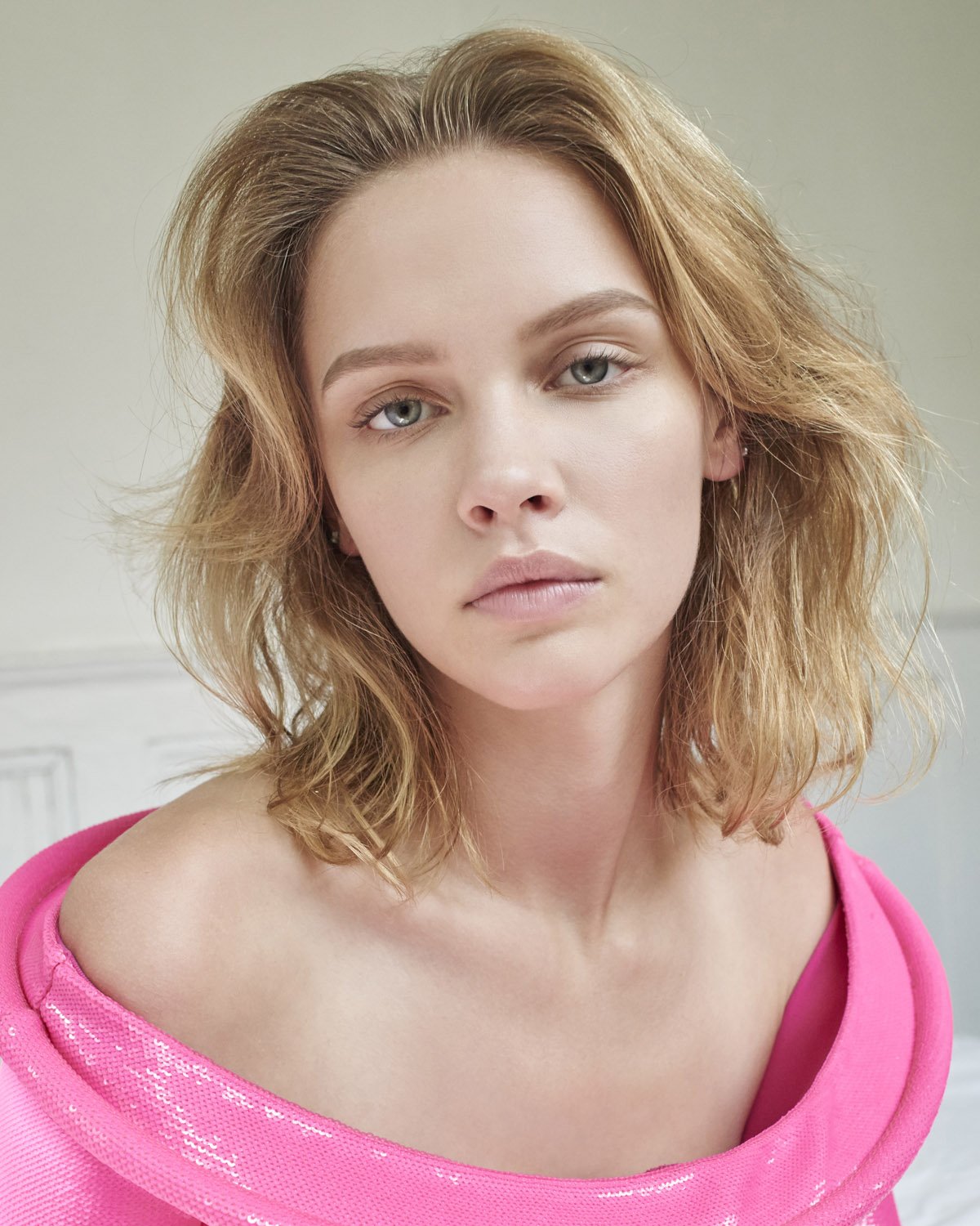 Portrait of a blonde woman wearing a vibrant pink off-shoulder garment against a plain interior background
