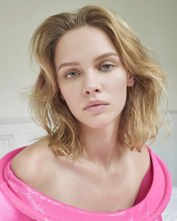 Portrait of a blonde woman wearing a vibrant pink off-shoulder garment against a plain interior background