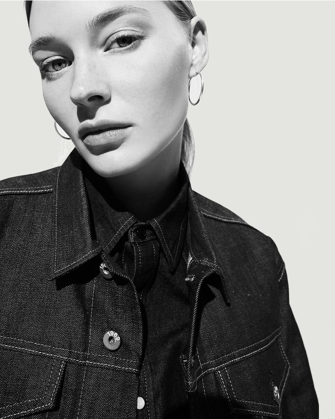Close-up portrait of a woman wearing a dark denim jacket and silver hoop earrings against a bright seamless background.