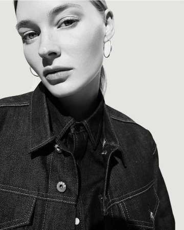 Close-up portrait of a woman wearing a dark denim jacket and silver hoop earrings against a bright seamless background.