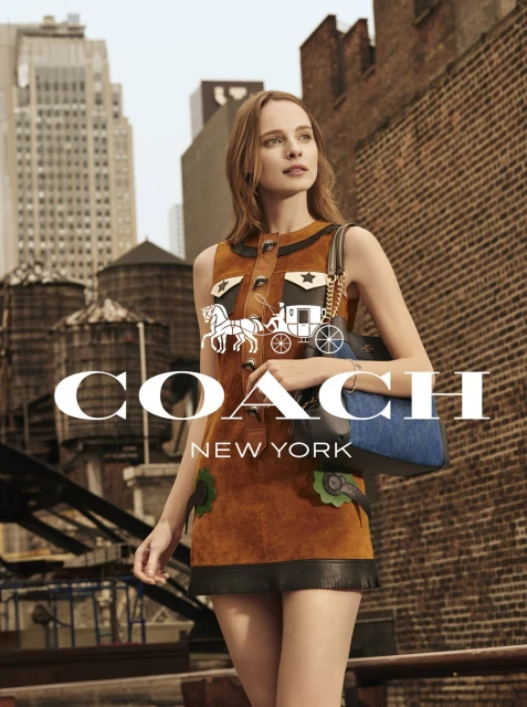 Fashion model poses wearing a brown minidress with a denim shoulder bag in an outdoor urban setting against a brick wall.