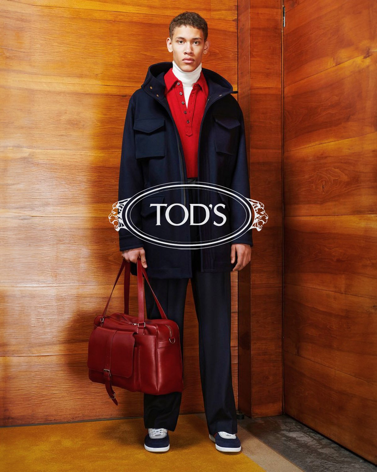 A dark-skinned male model poses in a blue coat and holds a red leather bag in a fashion shot against a wooden background indoors.