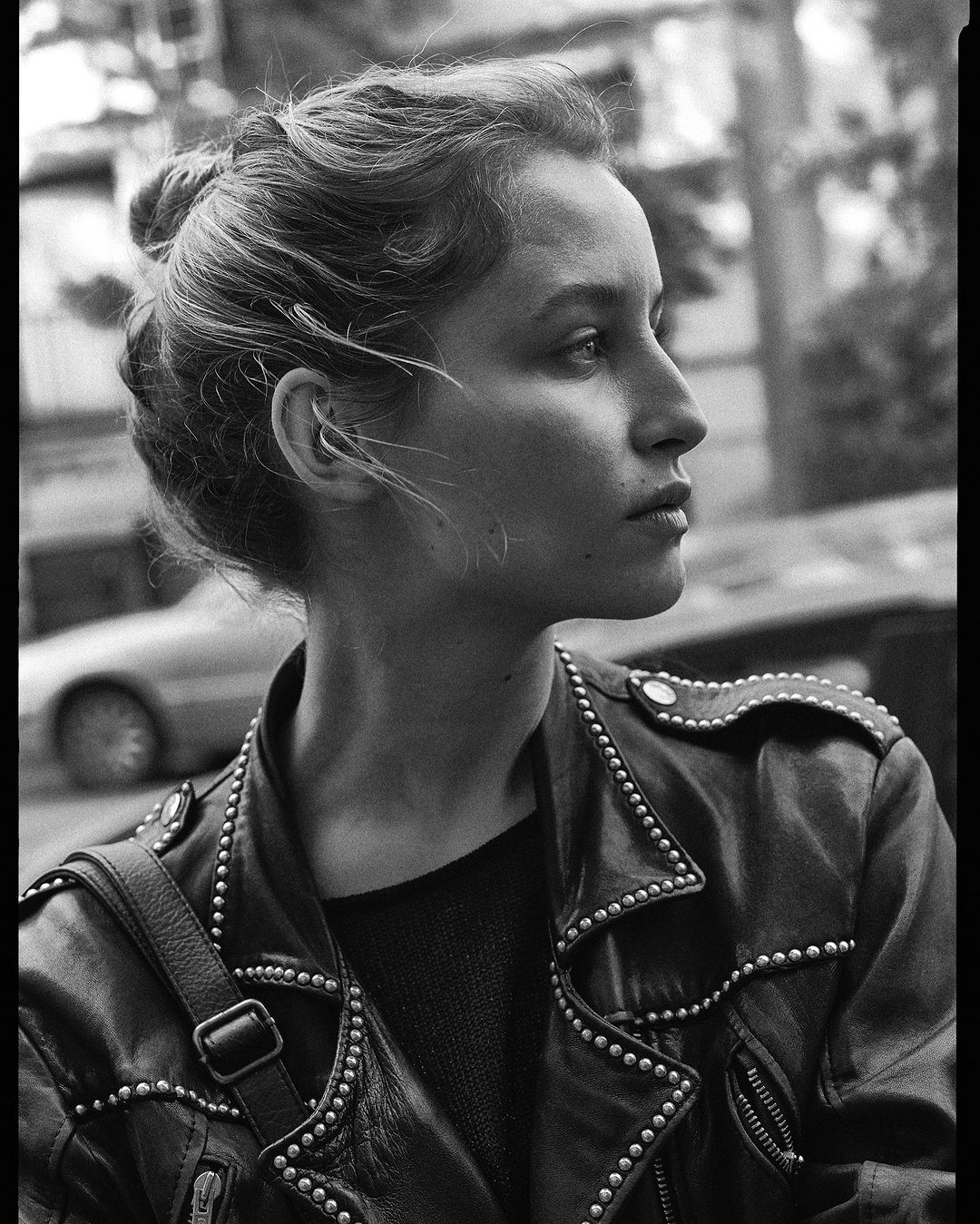 Monochrome portrait of a young woman with her hair up, wearing a studded leather jacket looking off to the side. Automotive background.