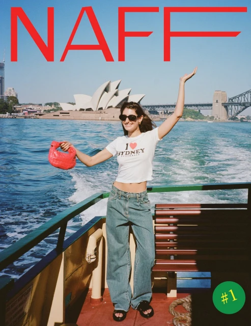 Woman on boat with Sydney Opera House and Harbour Bridge in background. She wears a white top, jeans, and holds a red purse.