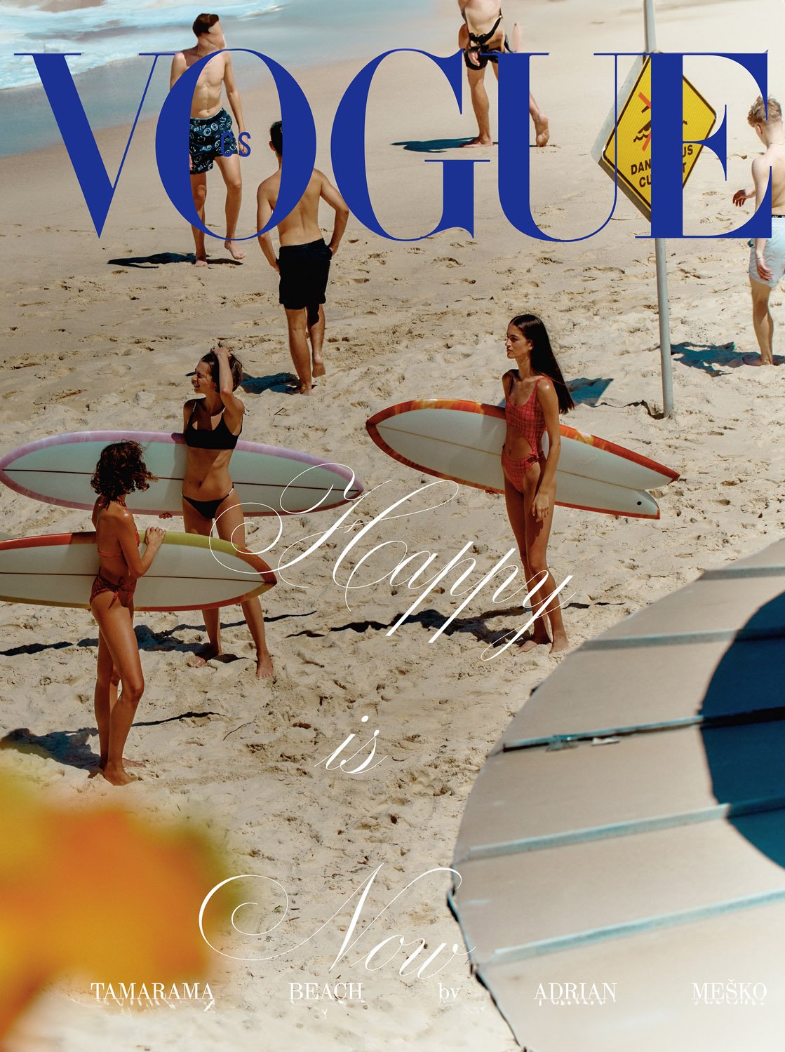 Vogue magazine cover on a beach with a group of women in swimsuits holding surfboards; men in swim trunks stand in the background.