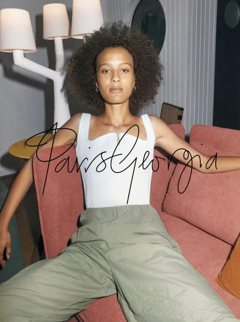 A young woman with curly hair poses while seated on an orange couch in front of a white floor lamp and striped wall.