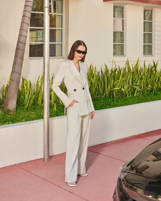 A woman in a checkered pantsuit and sunglasses poses on a pink sidewalk in front of a building on a sunny day in an urban setting.