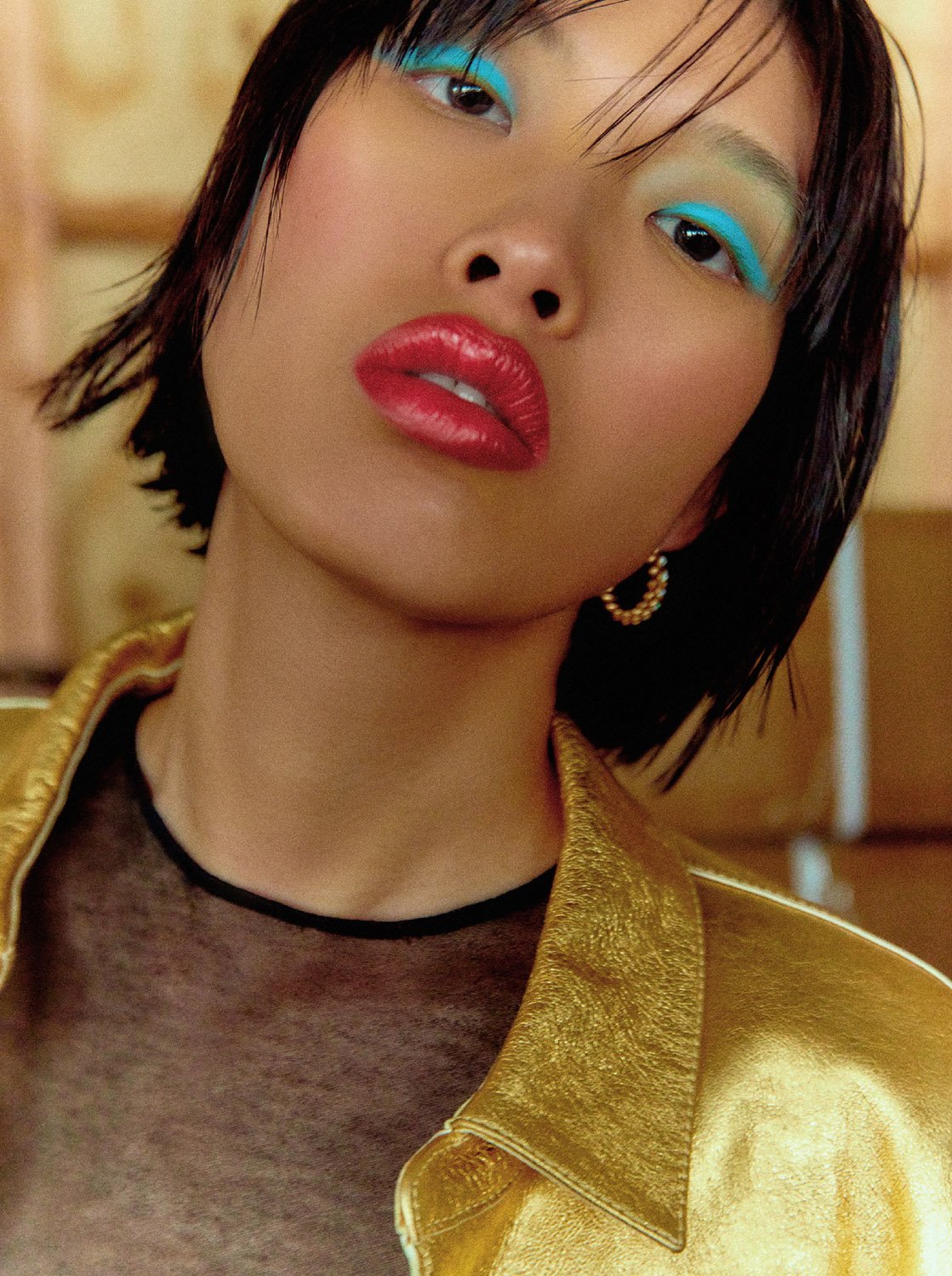 Close-up portrait of a model with vibrant blue eyeshadow and red glossy lips wearing a metallic gold jacket.