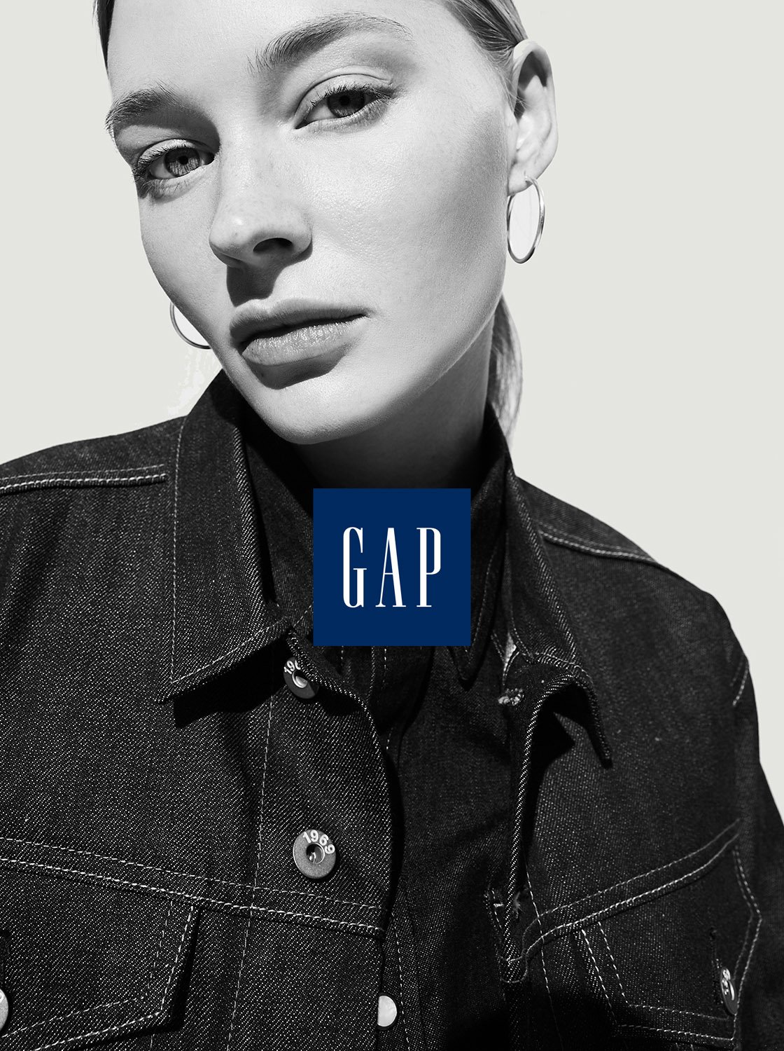 Close-up studio shot of a young woman with fair skin wearing denim jacket and hoop earrings, featuring the GAP logo in navy blue.