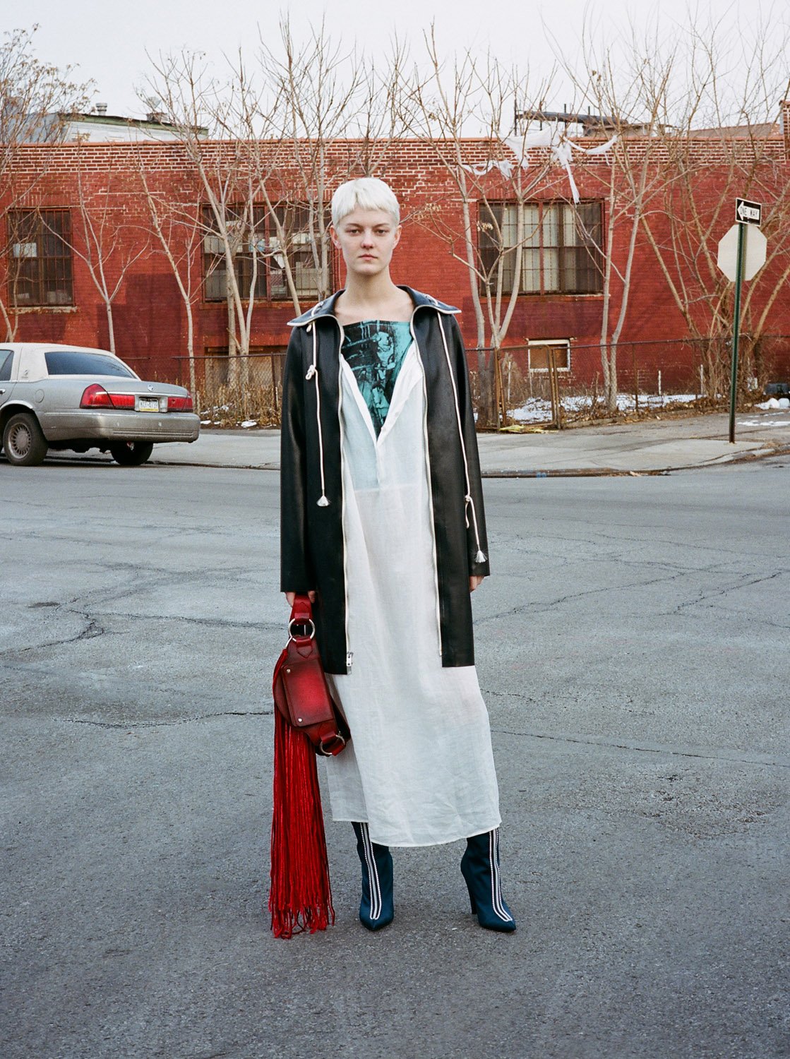 A woman with short platinum hair stands on an urban street wearing a white dress, black jacket, and boots. She carries a fringed purse.