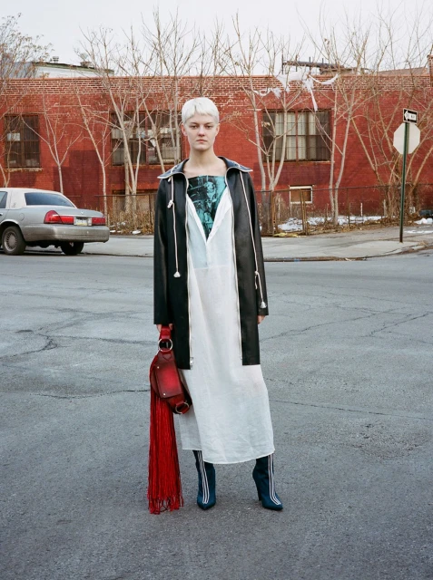 A woman with short platinum hair stands on an urban street wearing a white dress, black jacket, and boots. She carries a fringed purse.