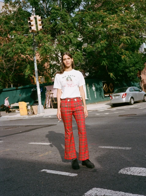 A young woman poses on a city street wearing a t-shirt and red plaid pants. There is a traffic light and building behind her.