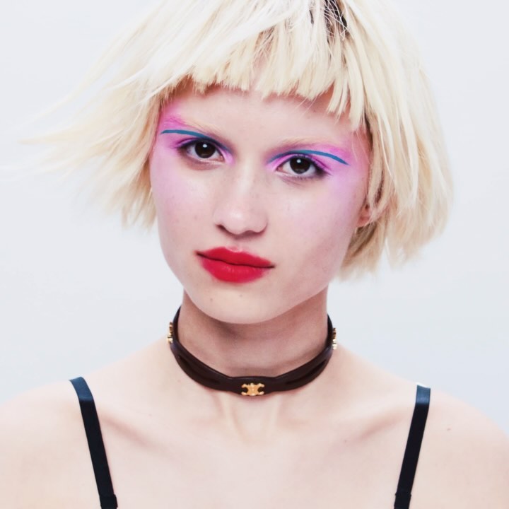 Close-up of a young woman with red lipstick, pink eyeshadow, and a black choker against a bright white background in a studio setting.