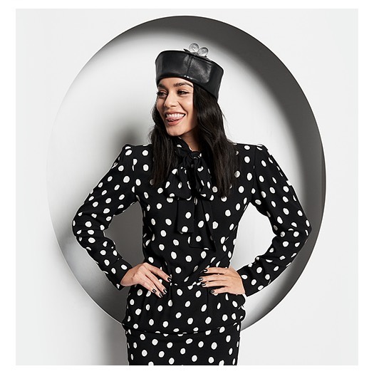 Smiling woman in polka dot suit and leather cap with wind-up key accent posing in front of white circular backdrop.