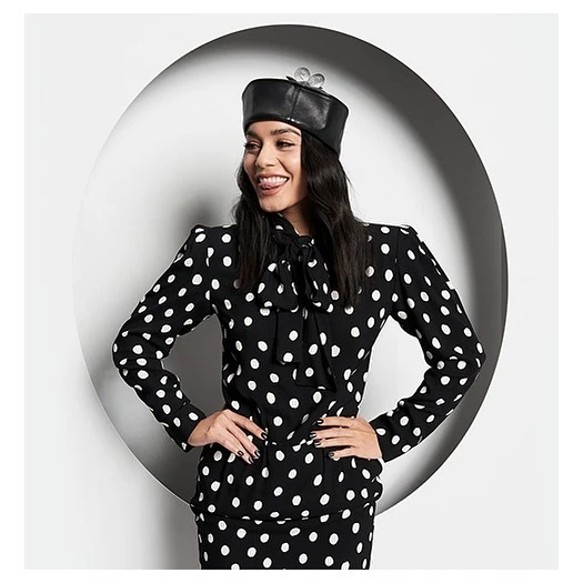 Smiling woman in polka dot suit and leather cap with wind-up key accent posing in front of white circular backdrop.