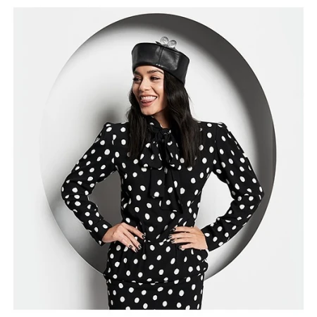 Smiling woman in polka dot suit and leather cap with wind-up key accent posing in front of white circular backdrop.