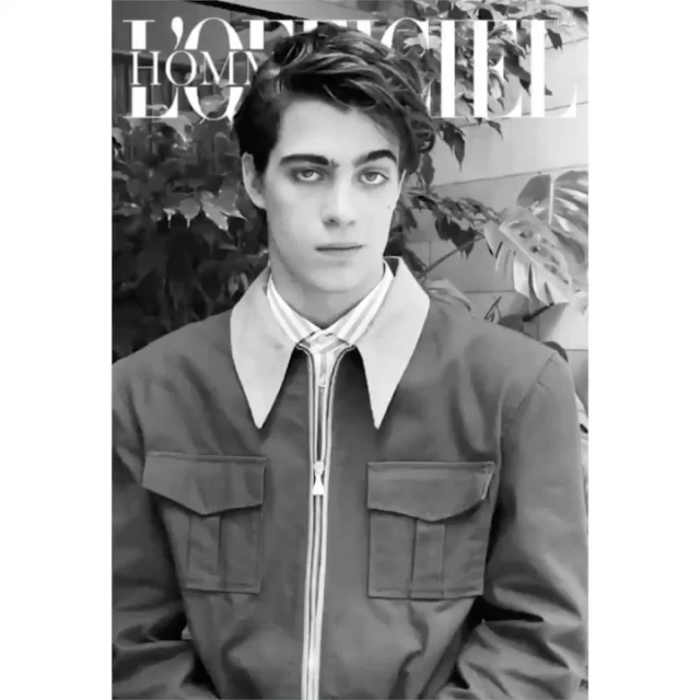 Monochrome portrait of a male model wearing a zip-up jacket, looking directly at the camera in front of a plant and a magazine cover.