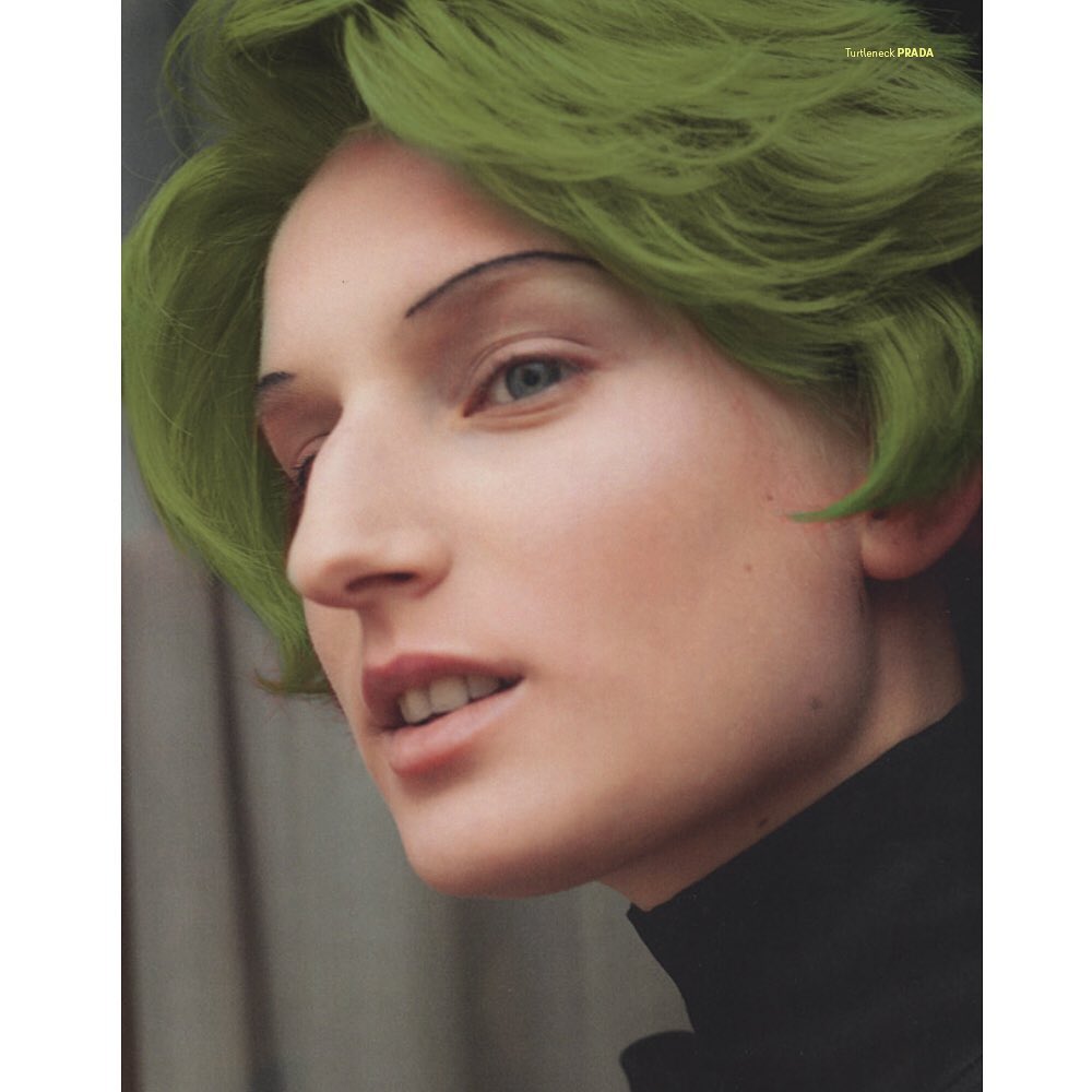Close-up portrait of a fair-skinned woman with short, wavy green hair, wearing a black turtleneck sweater against a grey background.