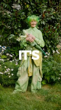 Senior woman with green hair, matching outfit, and an animal sculpture in a garden setting with green foliage and white flowers.