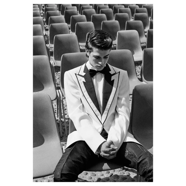 Monochrome portrait of a young adult male in a white tuxedo seated among rows of empty chairs in an outdoor setting.