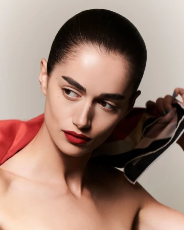 Close-up portrait of a woman with slicked-back dark hair wearing red lipstick and holding a patterned silk scarf.