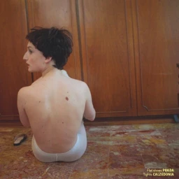 A woman with short dark hair viewed from behind, seated on a reddish marble floor against wooden cabinets.