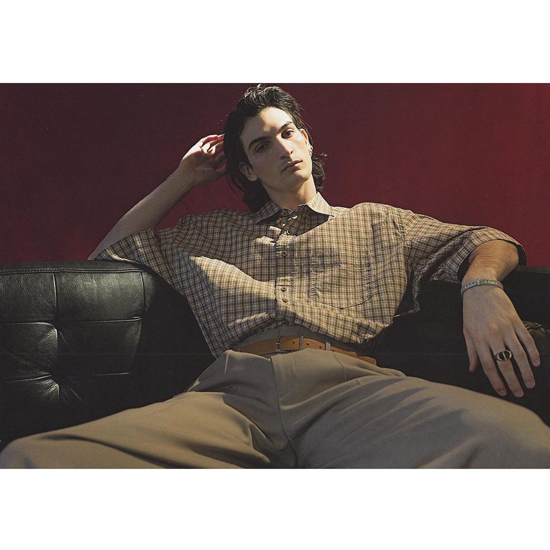 Young man with dark hair wearing a plaid short-sleeve shirt and trousers seated on a black leather sofa.