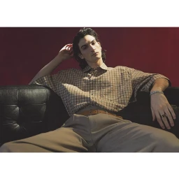 Young man with dark hair wearing a plaid short-sleeve shirt and trousers seated on a black leather sofa.