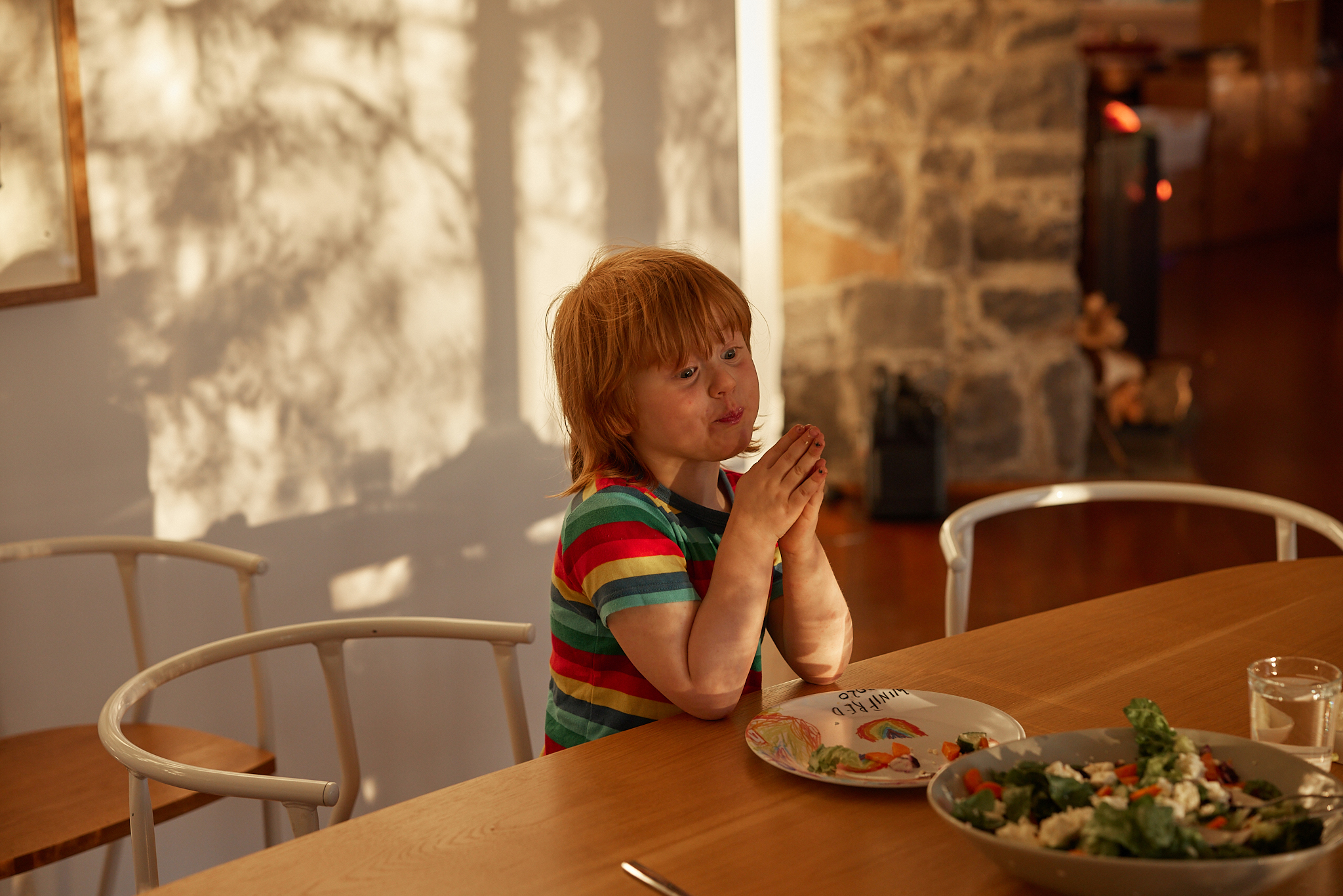 A child with red hair sits at a light wood table with a salad and plate in front of them in a brightly lit home interior.
