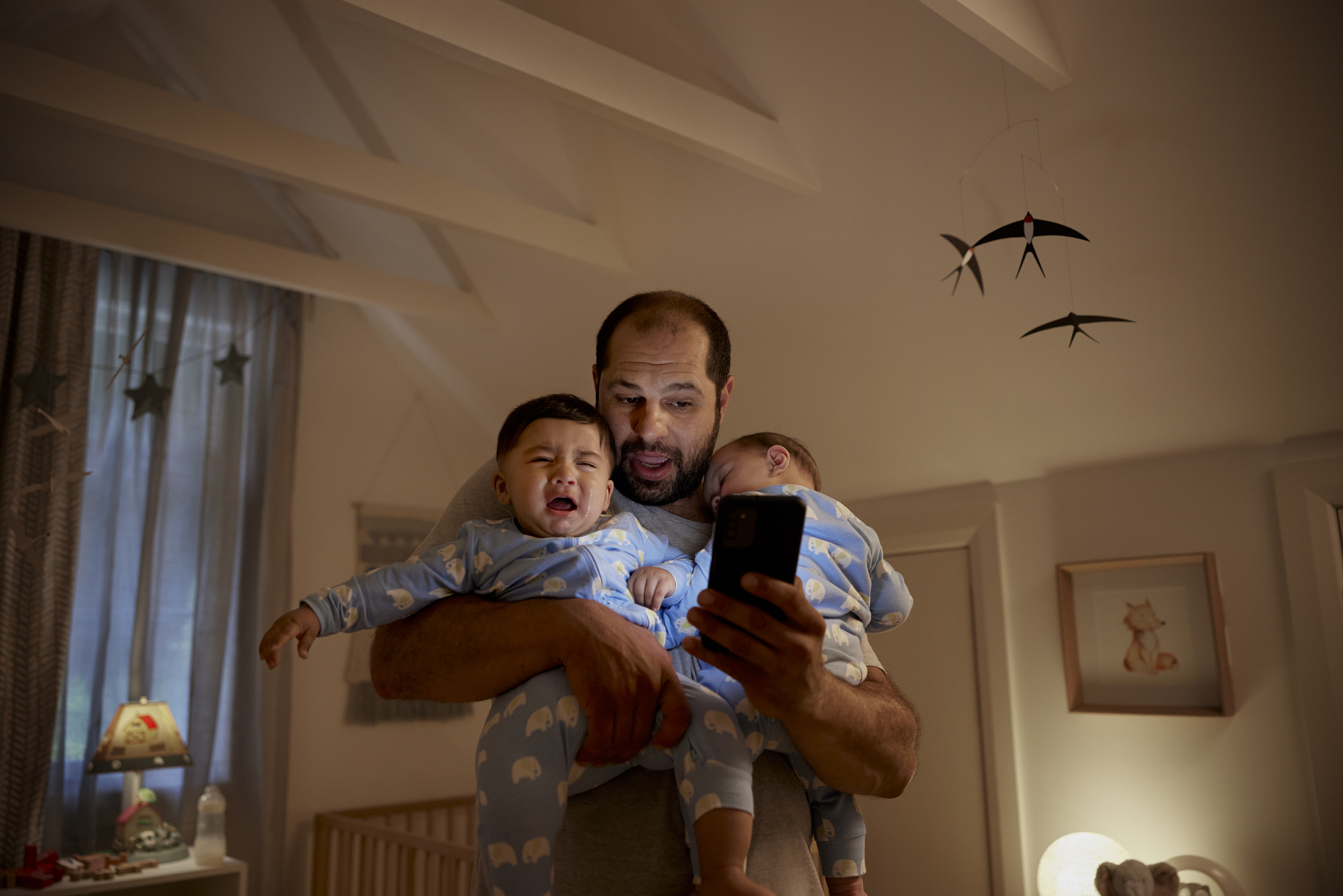 Father holding crying baby and another sleeping baby, looking at a smartphone in a dimly lit room with nursery decor.