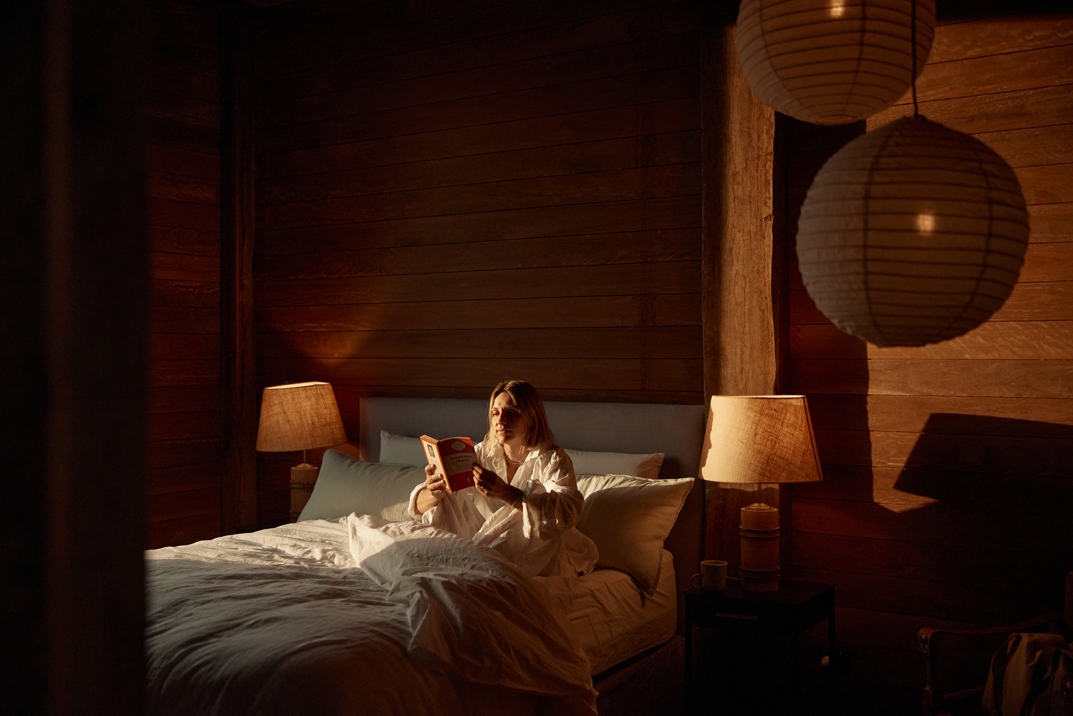 Adult female reading a book in bed inside a rustic cabin bedroom with soft lighting and round hanging paper lanterns.