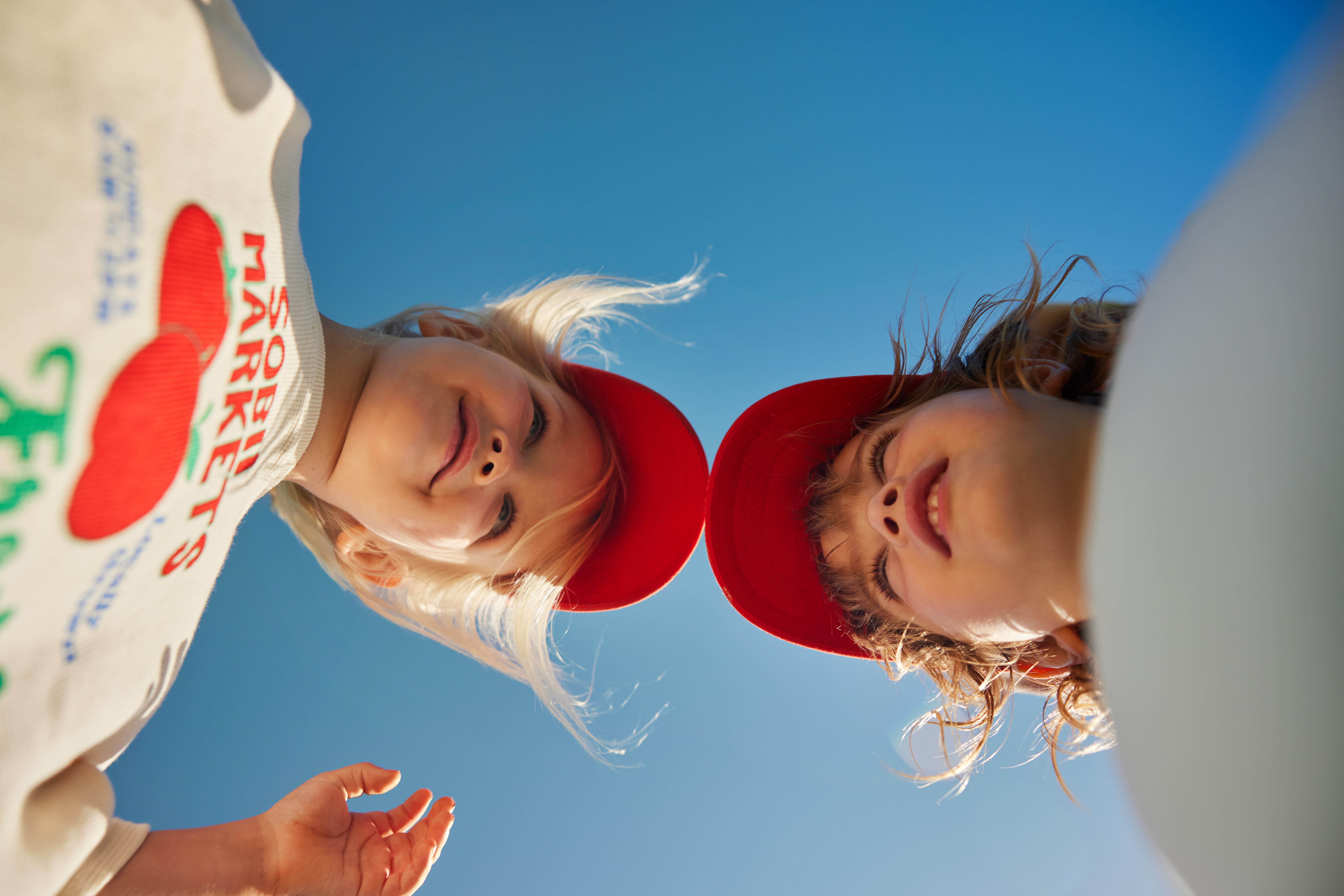 Two young children wearing red caps and a light-colored printed sweatshirt look down at the camera against a clear blue sky background.