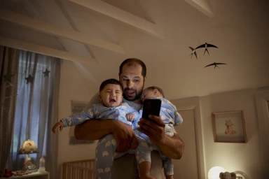 Man holding two crying toddlers in matching blue pajamas while looking at a smartphone in a nursery interior.