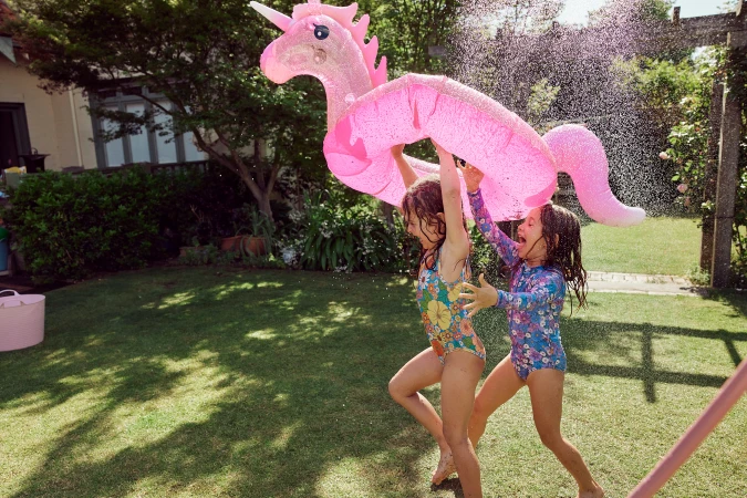 Two young girls playing in a sunny backyard while holding a large pink glittery unicorn pool float under a water spray