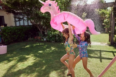 Two young girls playing in a sunny backyard while holding a large pink glittery unicorn pool float under a water spray